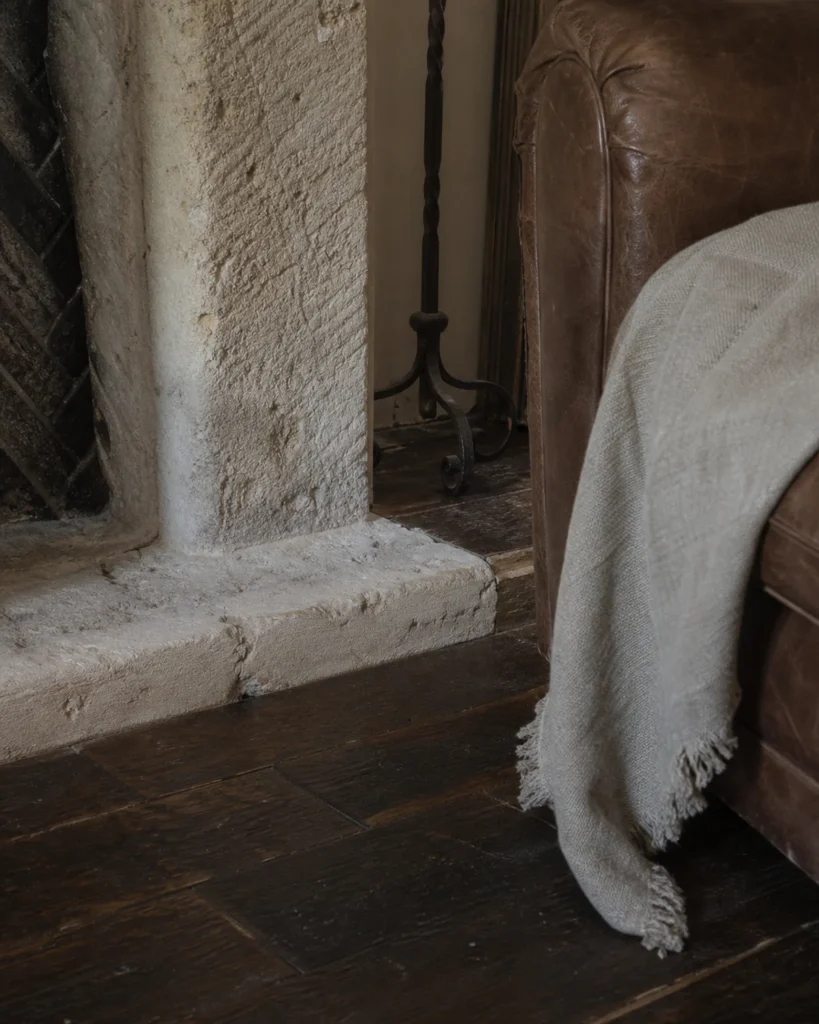 Close view of an irregular pale fireplace texture, dark wood-toned floor, worn brown leather-like upholstery and a plain flax-look textile.